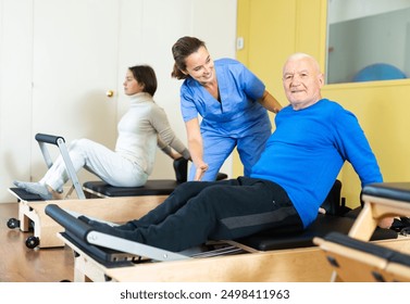 Professional female rehabilitation physician controlling movements of senior man doing exercises on pilates reformer - Powered by Shutterstock