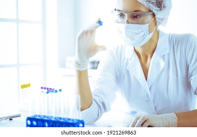 Professional female scientist in protective eyeglasses researching tube with reagents in sunny laboratory toned in blue. Medicine and science researching