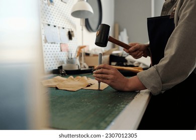 Professional female tanner keeping hammer over small steel shaft while pressing it down leather piece during work in craftshop or studio - Powered by Shutterstock