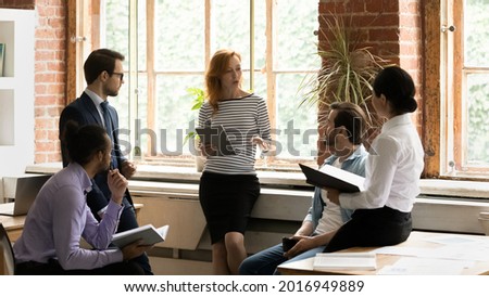 Professional female team leader holding digital computer tablet in hands, presenting business ideas to mixed race colleagues. Concentrated diverse coworkers discussing project development strategy.