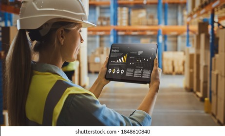 Professional Female Worker Wearing Hard Hat Uses Digital Tablet Computer With Screen Showing Analysis Software In The Retail Warehouse Full Of Shelves With Goods. Over The Shoulder View