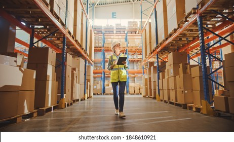 Professional Female Worker Wearing Hard Hat Checks Stock And Inventory With Digital Tablet Computer Walks In The Retail Warehouse Full Of Shelves With Goods. Working In Delivery, Distribution Center