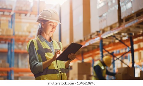 Professional Female Worker Wearing Hard Hat Checks Stock and Inventory with Digital Tablet Computer in the Retail Warehouse full of Shelves with Goods. Working in Logistics, Distribution Center