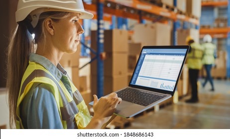 Professional Female Worker Wearing Hard Hat Holds Laptop Computer With Screen Showing Inventory Checking Software In The Retail Warehouse Full Of Shelves With Goods. Over The Shoulder Side View