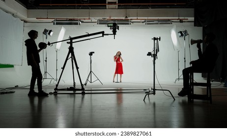 Professional filming pavilion with a white cyclorama. Shooting of a girl in a red dress who sings into a retro microphone. Director, Cameraman and crew in Backstage. - Powered by Shutterstock