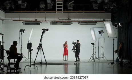 Professional filming pavilion with a white cyclorama. Shooting of a girl in a red dress who sings into a retro microphone. Director, Cameraman and crew in Backstage. - Powered by Shutterstock