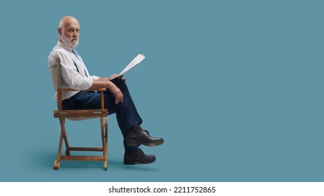 Professional Filmmaker Sitting On The Director's Chair And Holding A Screenplay, He Is Looking At Camera