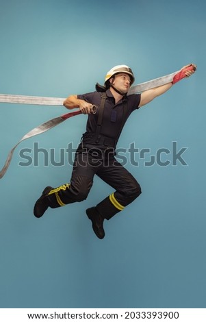 Professional firefighter. Vertical portrait of young man in uniform with water hose flying, jumping over blue studio background. Concept of different professions, unusual people and their emotions.