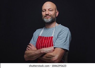 Professional Fishmonger Or Butcher In Grey T-shirt And Red And White Stripe Classic Apron On A Black Background. Bold Male With Grey Beard In His 40s, Slim Body Type. Hands Crossed Smile Expression