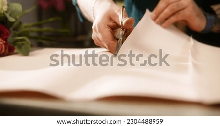 Professional florist puts on table wrapping paper and cuts it using scissors. Process of work in flower shop. Beautiful tied bouquet lies near. Concept of retail floral business and entrepreneurship.