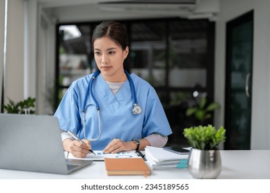 A professional and focused Asian female doctor in scrubs is working and reading medical research on her laptop in her office at a hospital.