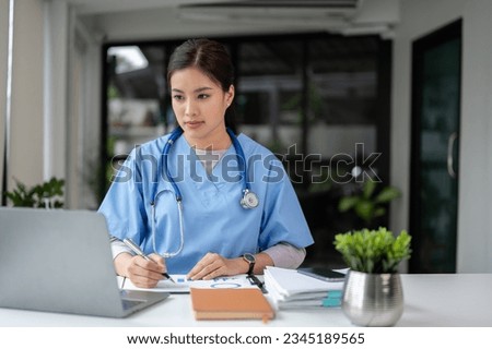 A professional and focused Asian female doctor in scrubs is working and reading medical research on her laptop in her office at a hospital.