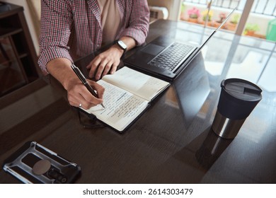 A professional freelancer taking notes for a startup plan at a desk with a laptop and coffee, showcasing focus and productivity in a home workspace, ideal for business and planning concepts.