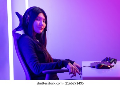 Professional Gamer. Young Asian Pretty Woman Sitting On Chair With Computer Pc In Living Room. Happy Female Professional Streamer Chinese Wearing Headphone Playing Game Online In Dark Room Neon Light