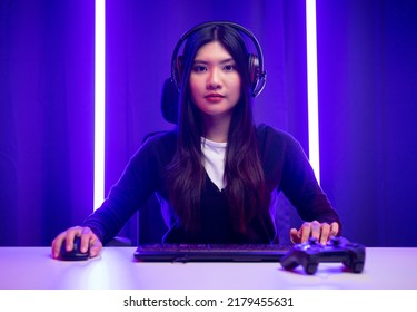 Professional gamer. Young asian pretty woman sitting on chair with computer pc in living room. Happy female Professional Streamer chinese wearing headphone playing game online in dark room neon light - Powered by Shutterstock