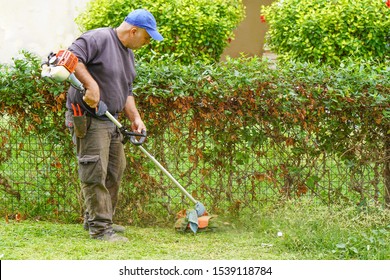Professional gardener using an edge trimmer in the city parck. Elderly man worker mowing lawn with grass trimmer outdoors on sunny day.                               
