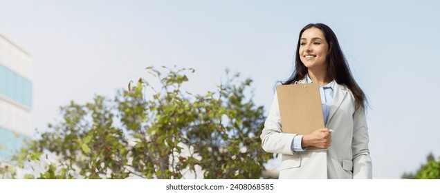 Professional glad caucasian young woman outdoors holding a clipboard, dressed smartly in a suit jacket, looking optimistically towards her future in the business world, panorama