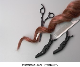 Professional Hairdresser Equipment On Gray Background. Scissors, Comb, Hairclip And Strand Of Curly Hair, Flat Lay