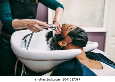 Professional Hairdresser Washing Hair Of A Middle Aged African American Woman With Protective Face Mask In Hair Salon. 