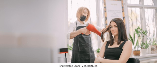 Professional hairdresser woman in an apron makes a hairstyle with a hair dryer to a young girl in a beauty salon. A young woman is sitting on the styling in the hairdresser's salon. - Powered by Shutterstock