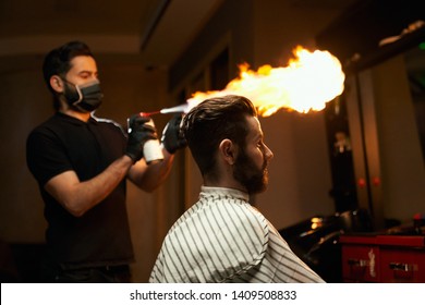 Professional Handsome Barber With Black Mask On Mouth Is Going To Make A Haircut With Fire Treatment To Male Bearded Client That Is Sitting In Chair And Covered By Striped Cape At Modern Barbershop