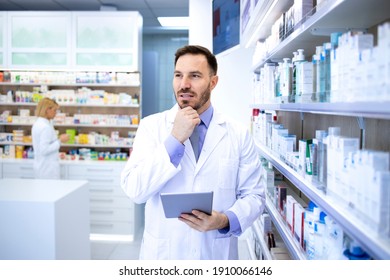 Professional Handsome Male Pharmacist In White Coat Holding Tablet And Thinking In Pharmacy Store Or Drugstore. Healthcare And Apothecary.