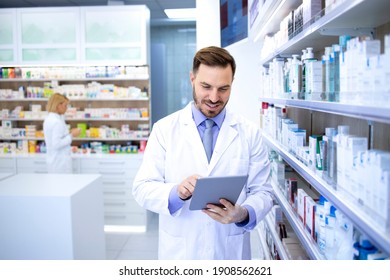 Professional Handsome Male Pharmacist Working In Pharmacy Store Or Drugstore. Checking Medicines On His Tablet Computer. Healthcare And Apothecary.