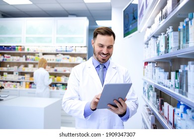 Professional Handsome Male Pharmacist Working In Pharmacy Store Or Drugstore. Checking Medicines On His Tablet Computer. Healthcare And Apothecary.