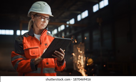 Professional Heavy Industry Engineer Worker Wearing Safety Uniform and Hard Hat Uses Tablet Computer. Serious Successful Female Industrial Specialist Standing in a Metal Manufacture Warehouse.