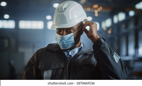 Professional Heavy Industry Engineer Worker Is Putting On Safety Face Mask In A Steel Factory. Black African American Industrial Specialist In Hard Hat Standing In A Metal Construction Manufacture.