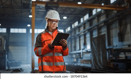 Professional Heavy Industry Engineer Worker Wearing Safety Uniform   Hard Hat  Using Tablet Computer  Serious Successful Female Industrial Specialist Walking in Metal Manufacture Warehouse 