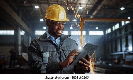 Professional Heavy Industry Engineer Worker Wearing Safety Uniform and Hard Hat Uses Tablet Computer. Smiling African American Industrial Specialist Standing in a Metal Construction Manufacture.