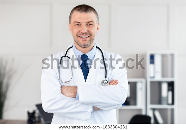 Professional Help. Portrait of smiling confident\
mature general practitioner standing with folded arms at his office\
in clinic and posing, looking at camera. Doctor wearing stethoscope\
and white coat
