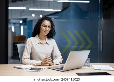 Professional Hispanic businesswoman smiling confidently while seated at her modern workspace, surrounded by digital devices in an office setting. - Powered by Shutterstock