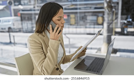 A professional hispanic woman multitasks with a laptop and phone while working at a modern office. - Powered by Shutterstock