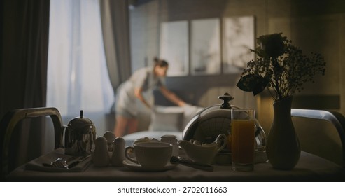 Professional Hotel Maid Opens Curtains on Windows and Makes Fresh, Neat Bed in Elegant Room. In Foreground Room Service Trolley With Breakfast. Comfort, Cleanliness, and High Quality Hotel Service.