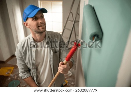 Professional house painter smiling while using a paint roller to refresh apartment walls with a light green color, enhancing the room's decor and creating a cheerful atmosphere