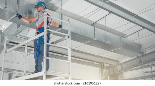 Professional HVAC Worker On A Aluminium Scaffolding Installing Air Duct Inside Newly Built Commercial Building Warehouse. Industrial Theme.