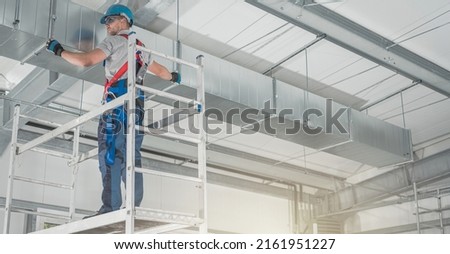 Professional HVAC Worker on a Aluminium Scaffolding Installing Air Duct Inside Newly Built Commercial Building Warehouse. Industrial Theme.