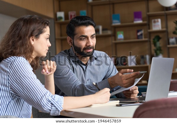 Professional indian teacher, executive or mentor\
helping latin student, new employee, teaching intern, explaining\
online job using laptop computer, talking, having teamwork\
discussion in\
office.