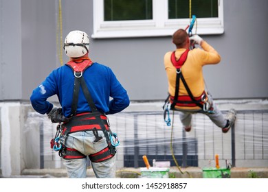 Professional Industrial Alpinist With Rigging Equipment, Hardhat And Safety Harness From The Back. Painter Hanging On Cable With Paint Buckets, Industrial Climber Repairing House Facade