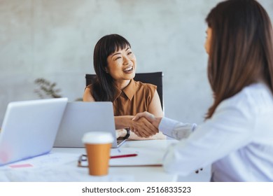 Professional interaction between two women shaking hands in an office setting, highlighting business collaboration and partnership. Laptops and coffee cups are visible in the workspace. - Powered by Shutterstock