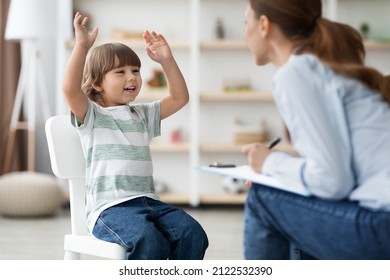 Professional Kids Psychotherapy. Happy Emotional Little Boy Talking To Therapist At Office, Sharing Funny Story To Specialist During Session