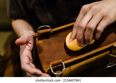 Professional leather care service concept. Closeup hand with yellow soft sponge cleaning stains on the surface of the vintage brown bag, depth of field. Polish waxing protection from dust and dirt.