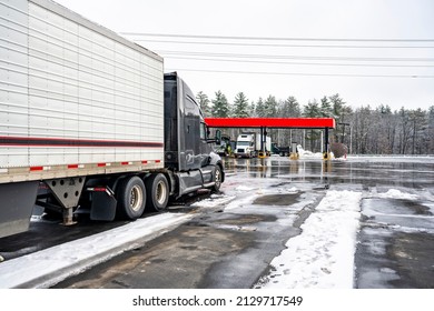 Professional Long Haul Black Big Rig Semi Truck With Refrigerator Semi Trailer Standing For Truck Driver Rest On The Winter Truck Stop Parking Lot With Snow And Ice And Covered Diesel Fuel Station