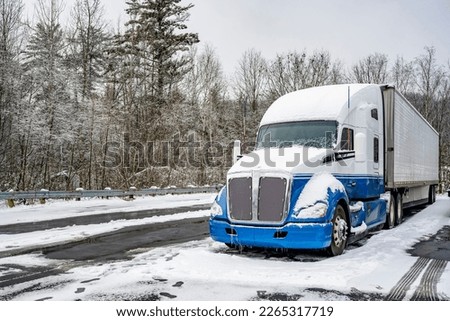 Professional long haul blue big rig semi truck tractor with refrigerator semi trailer standing for truck driver rest on the winter truck stop parking lot with snow and ice and trees on the background