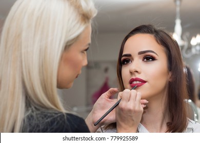Professional make-up artist applying bright red lipstick on beautiful girl using special lip brush.