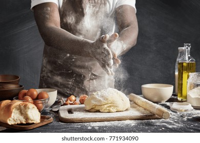 Professional male cook with dark skin, wears apron, sprinkles dough with flour, preapares or bakes bread at kitchen table, has dirty uniform, isolated over black chalk background. Baking concept - Powered by Shutterstock