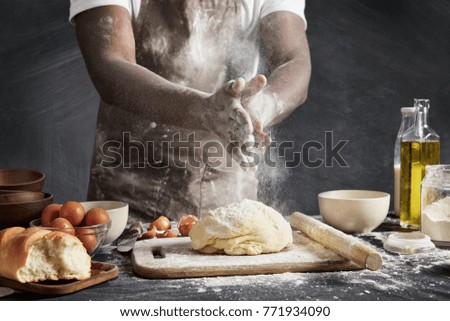 Professional male cook with dark skin, wears apron, sprinkles dough with flour, preapares or bakes bread at kitchen table, has dirty uniform, isolated over black chalk background. Baking concept