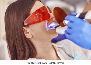 Professional male doctor uses ultraviolet lamp after making a filling on a tooth for joyful happy caucasian woman in modern dental cabinet - Powered by Shutterstock